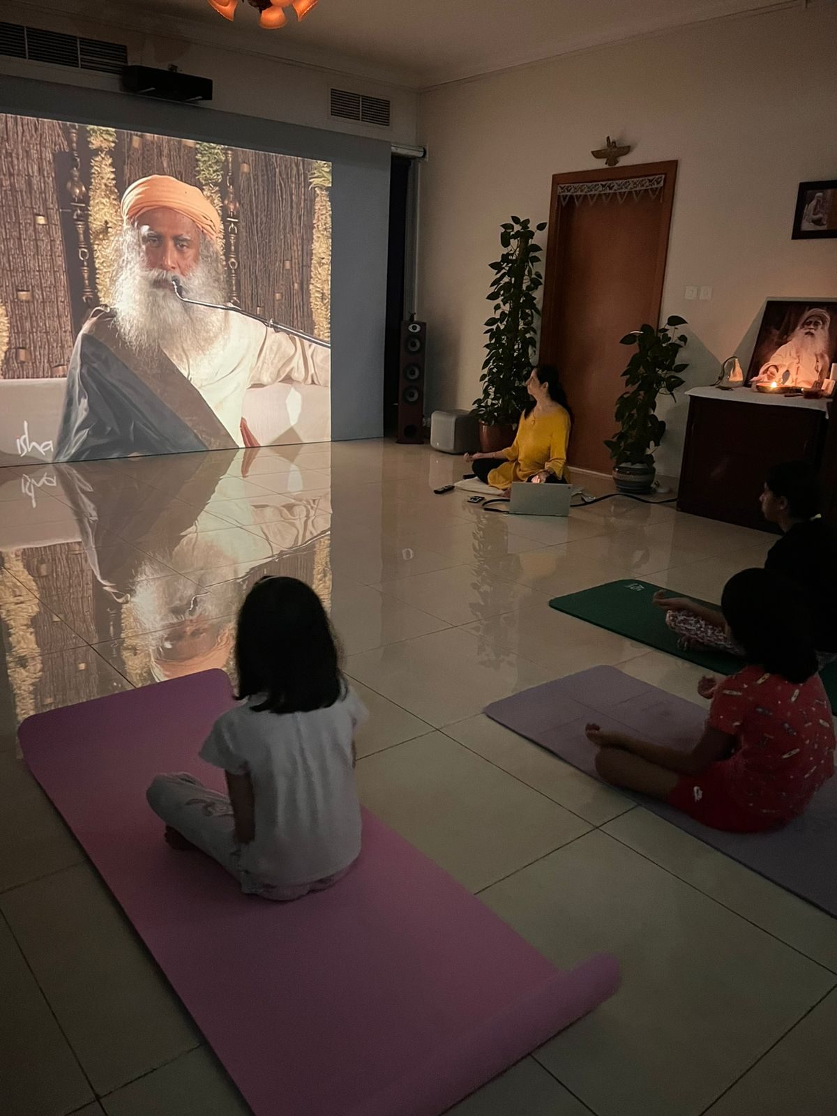 Children seated on mats watching Sadhguru on the studio screen with the lights dimmed