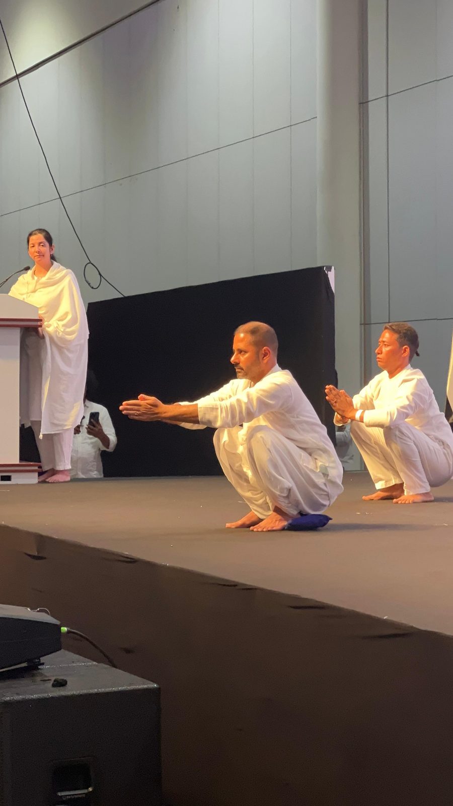 Persis on stage guiding two practitioners in a squat at International Yoga Day 2025
