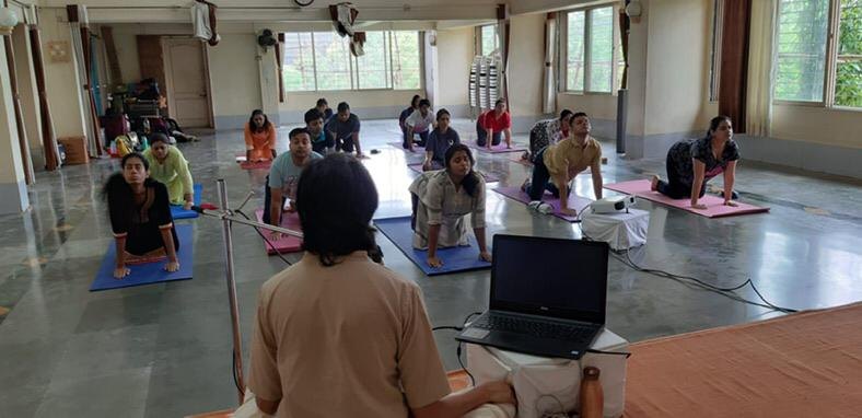 A room of adult students on mats facing Persis, following the practice she is guiding