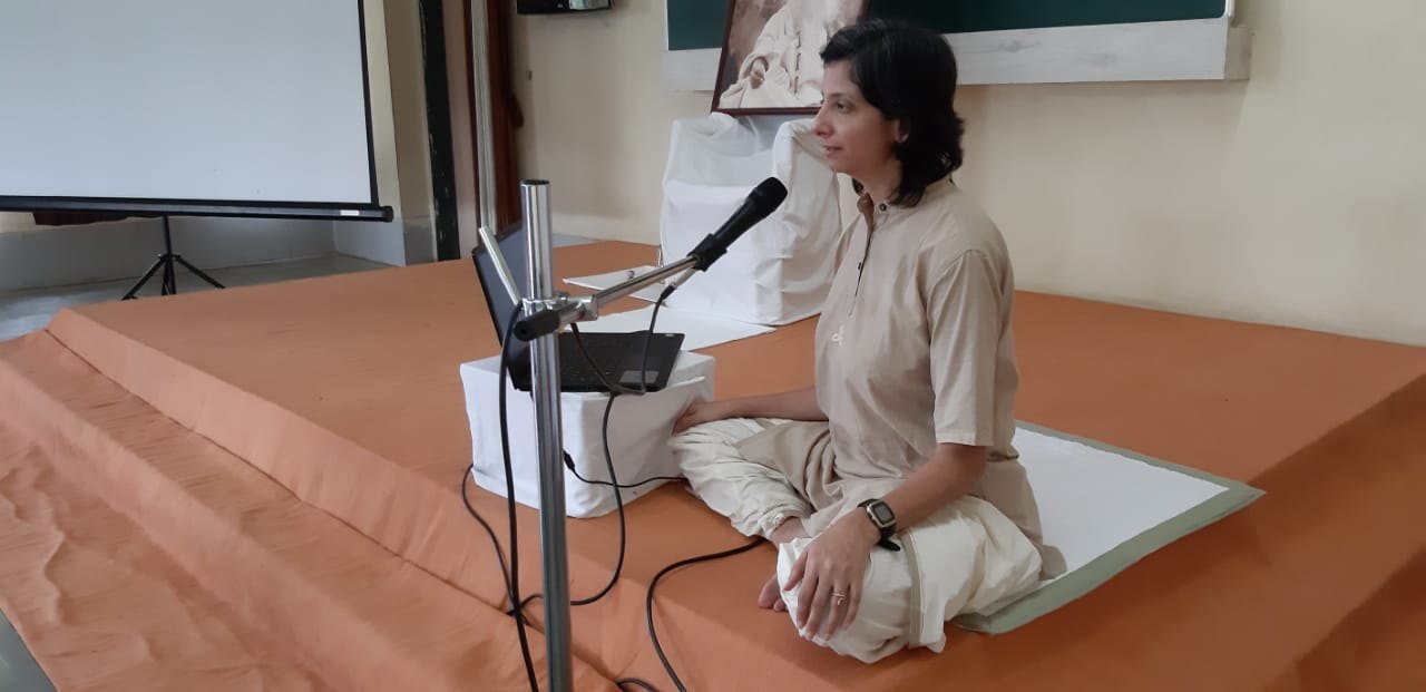 Persis seated on a low platform with a microphone in front of her, leading a session