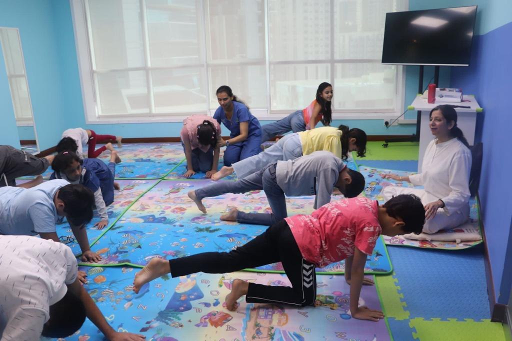 Persis seated at the side of a bright playroom guiding a group of children through a posture, a therapist helping one of them