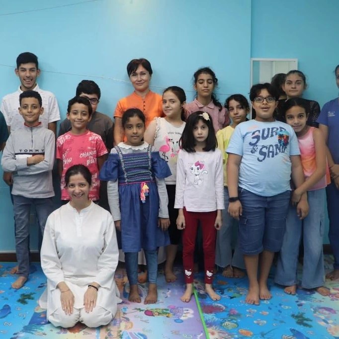 Persis kneeling in white among the children and staff of the centre after a session